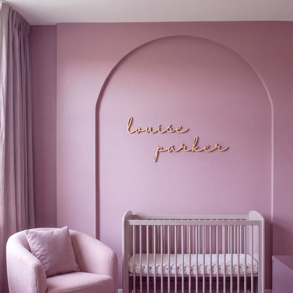 Cursive wood name sign above a crib in a pink nursery with an arched wall feature