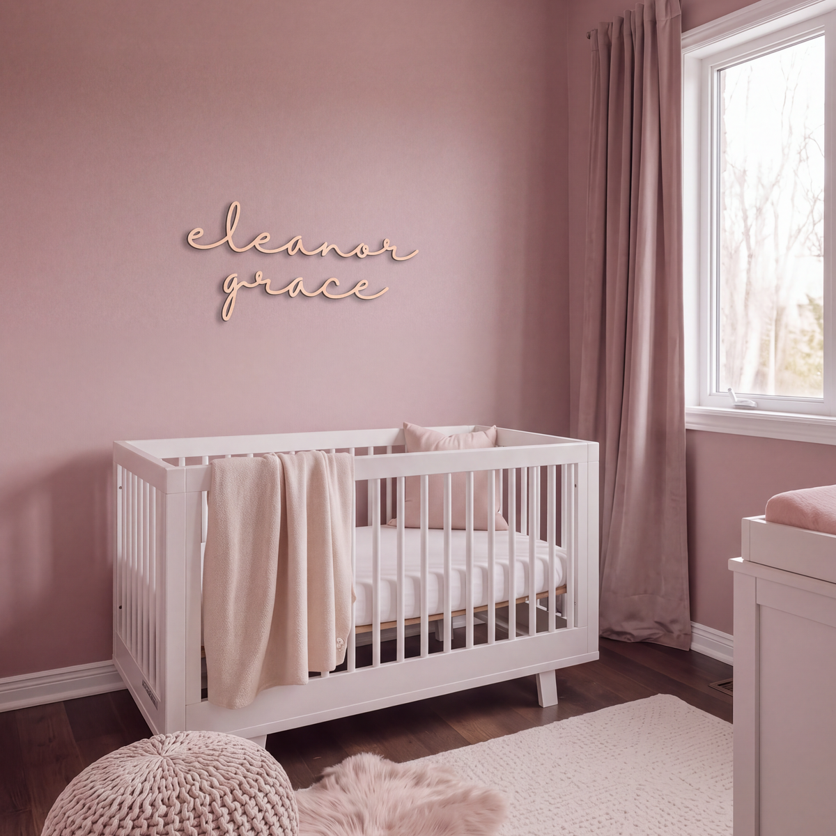 Natural wood cursive name sign in a pink nursery with a white crib and knitted pouffe
