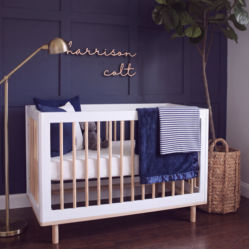Natural birch basswood cursive name sign on a navy panelled nursery wall above a white and wood crib