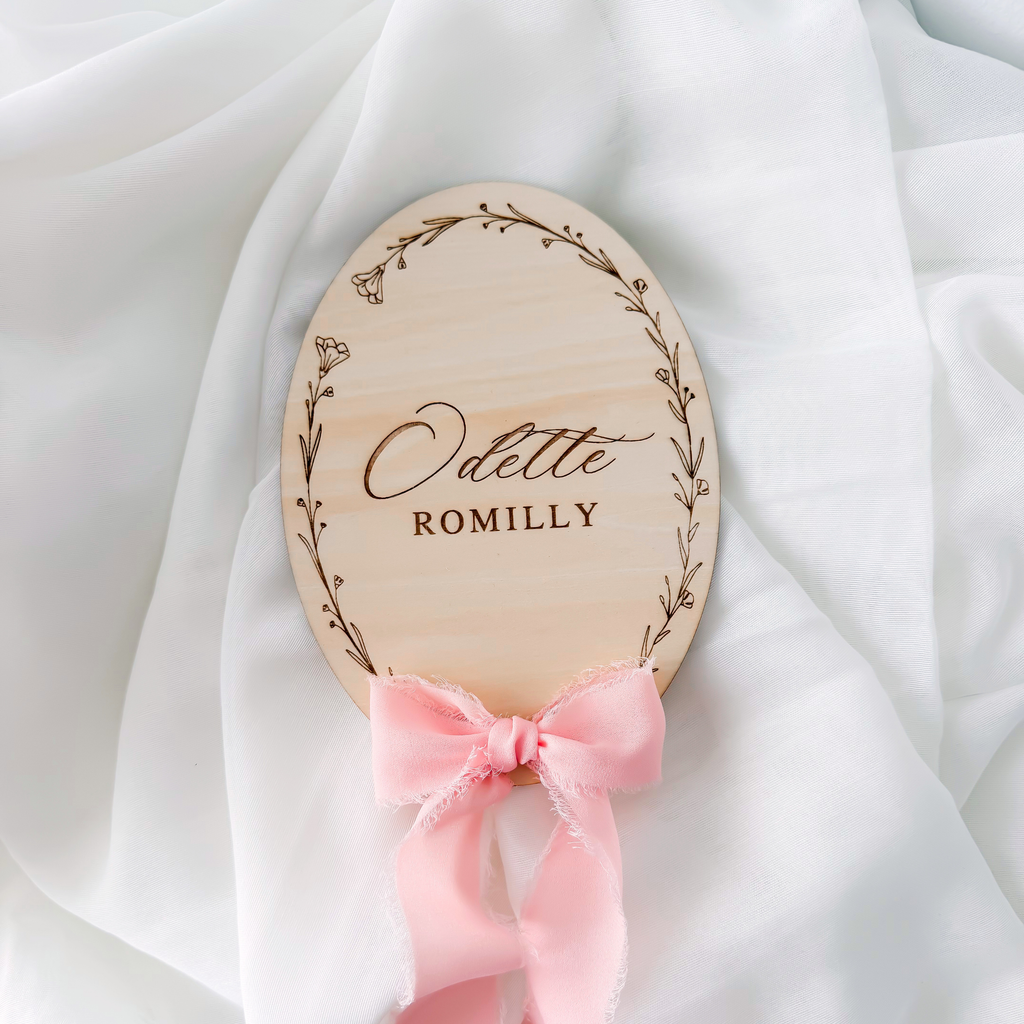 Floral wreath baby name sign lying on white fabric with a pink silk ribbon