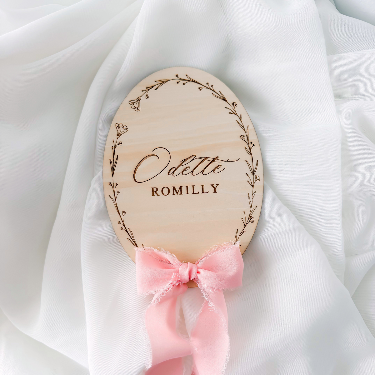 Floral wreath baby name sign lying on white fabric with a pink silk ribbon