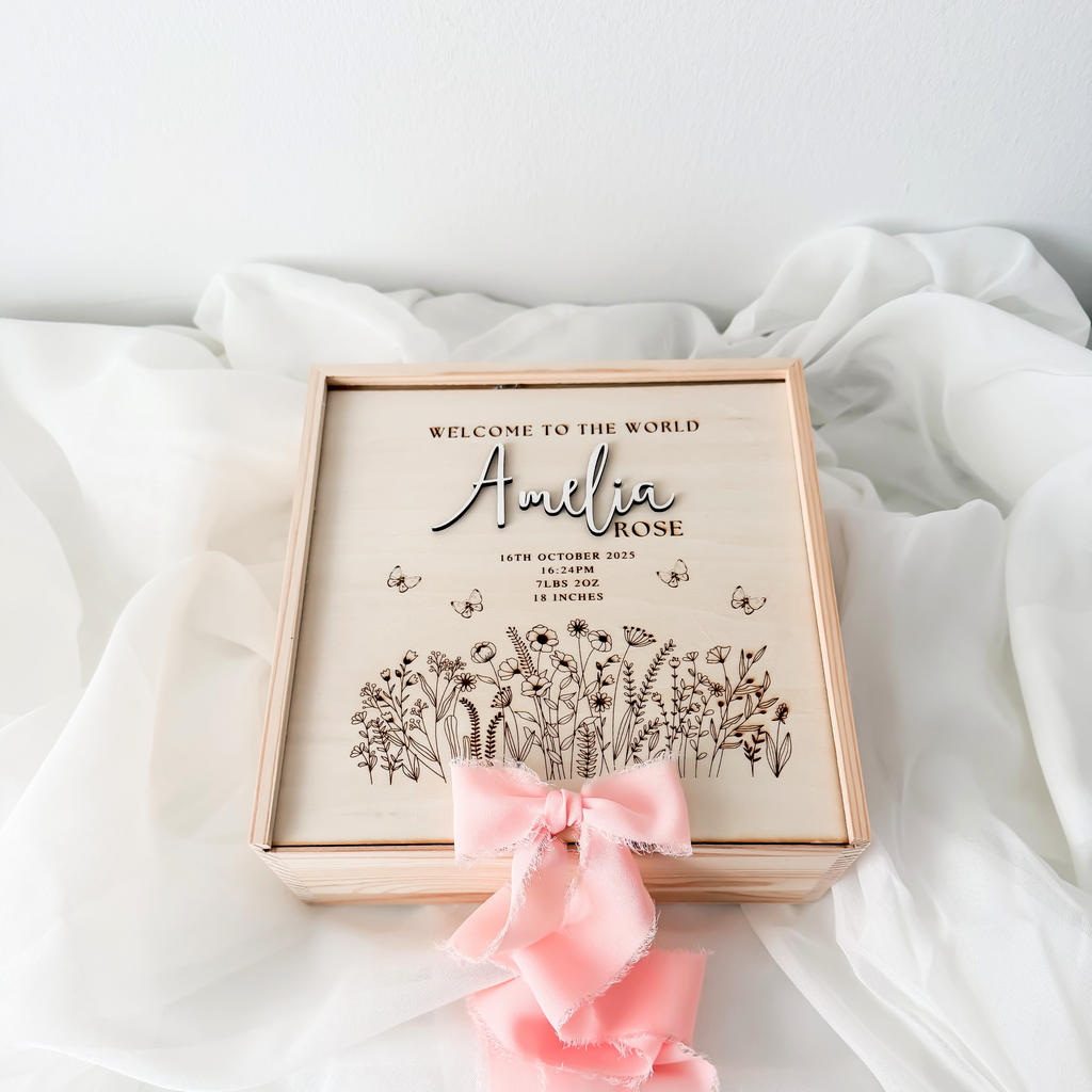 Wooden baby keepsake box with wildflower engraving and a pink silk ribbon, styled on white fabric