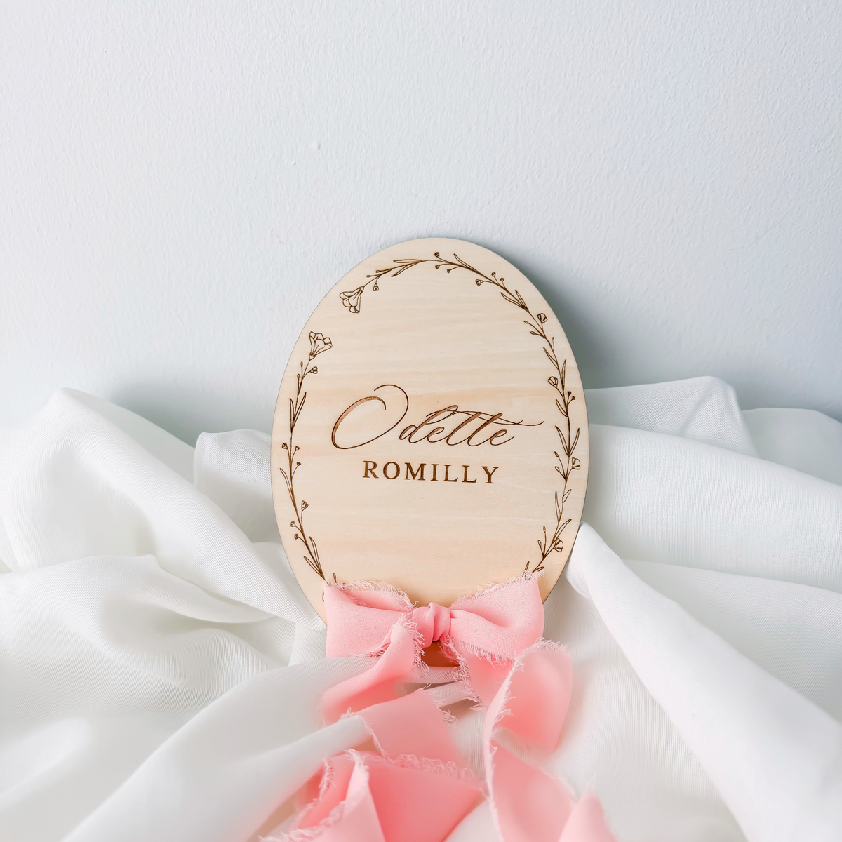 Oval birch basswood name sign with an engraved wildflower wreath and a pink silk ribbon bow