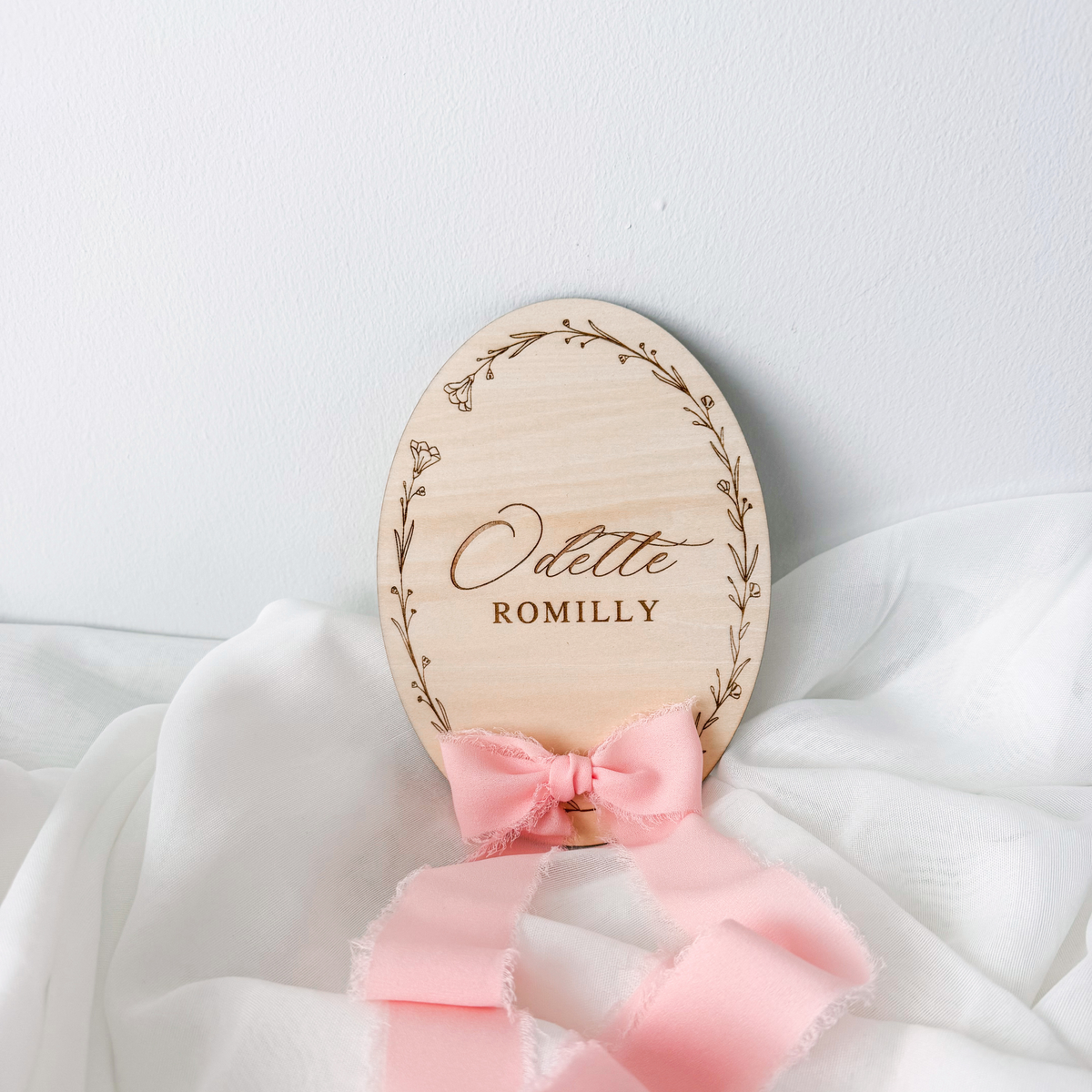 Floral wreath baby name sign engraved Odette Romilly, propped with a pink silk ribbon