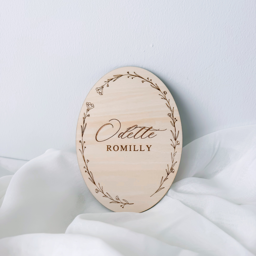 Oval floral wreath name sign leaning against a wall on white fabric, without a ribbon