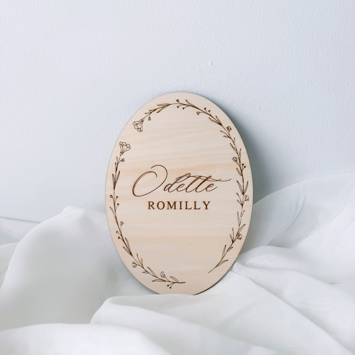 Oval floral wreath name sign leaning against a wall on white fabric, without a ribbon